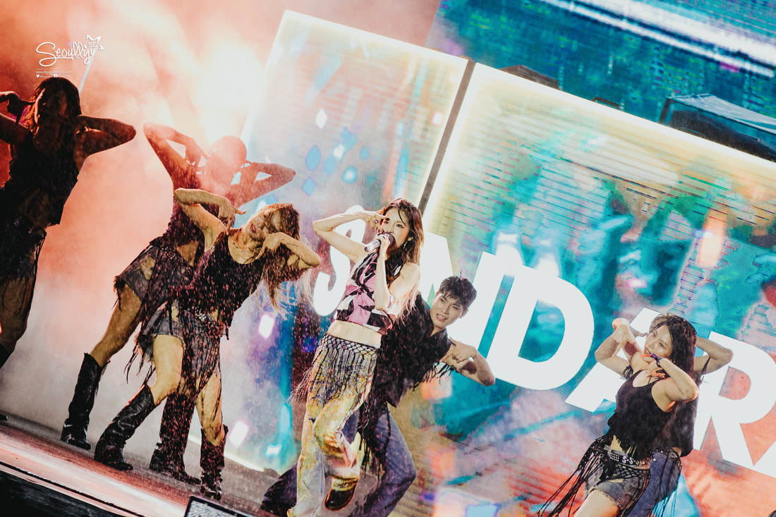 A dynamic performance with a singer at the center, surrounded by dancers, against a vibrant backdrop with colorful lighting. The dancers are dressed in edgy outfits, creating a lively atmosphere.