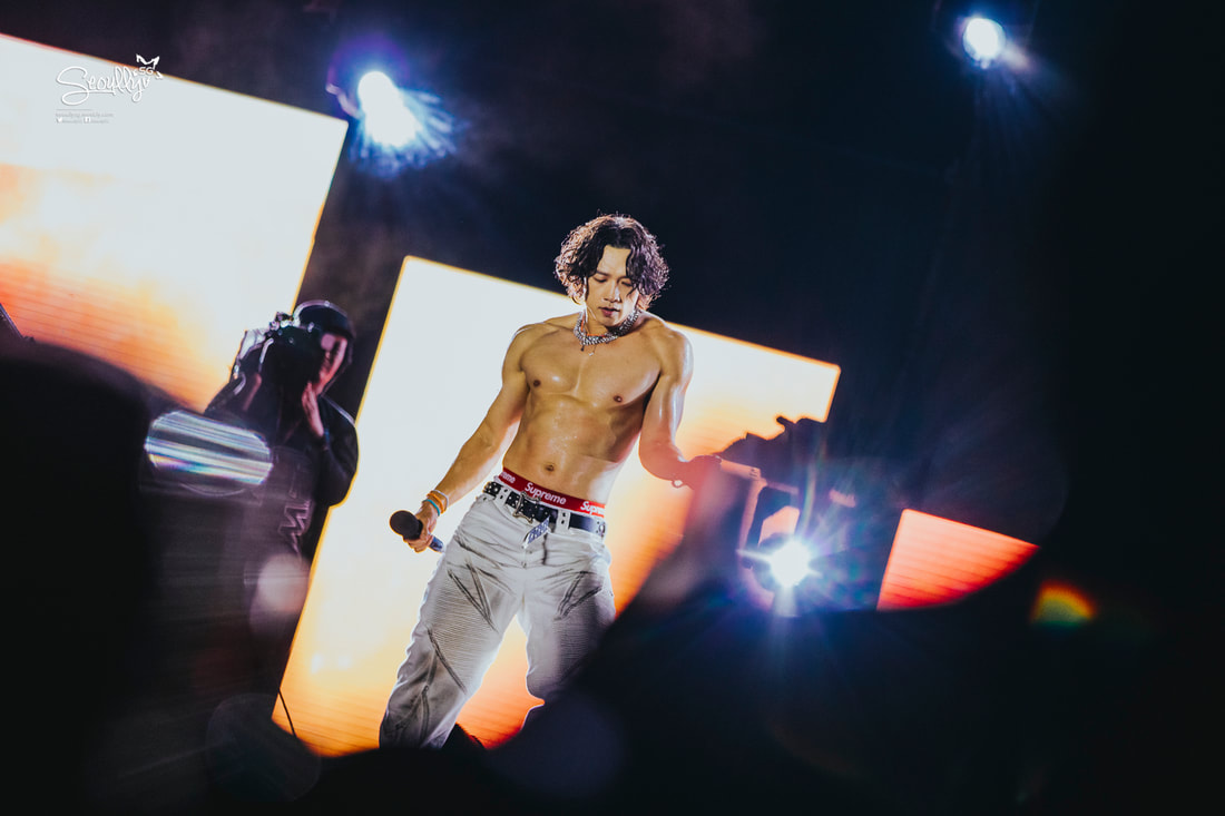 A male performer with curly hair is singing on stage, wearing no shirt and light-colored pants, with a microphone in hand. Bright stage lights illuminate the scene.