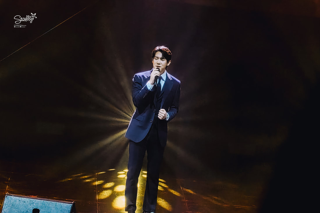 A performer in a formal suit singing passionately on stage, illuminated by dramatic lighting.