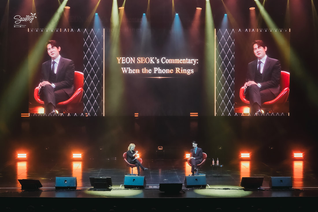 A stage setting featuring two speakers seated in red chairs, with a digital screen displaying 'YEON SEOK's Commentary: When the Phone Rings.' The atmosphere is enhanced by colorful stage lighting.