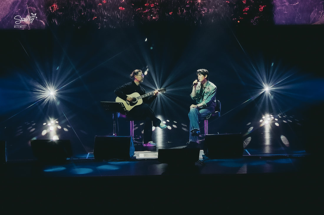 Two musicians perform on stage, one playing an acoustic guitar and the other singing into a microphone, surrounded by colorful stage lights.