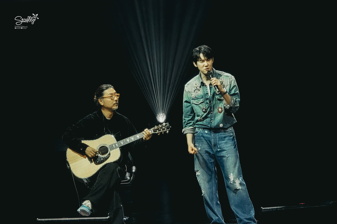 A singer performing on stage while holding a microphone, accompanied by a guitarist sitting with an acoustic guitar. Both are dressed in stylish outfits, with spotlight effects in the background.