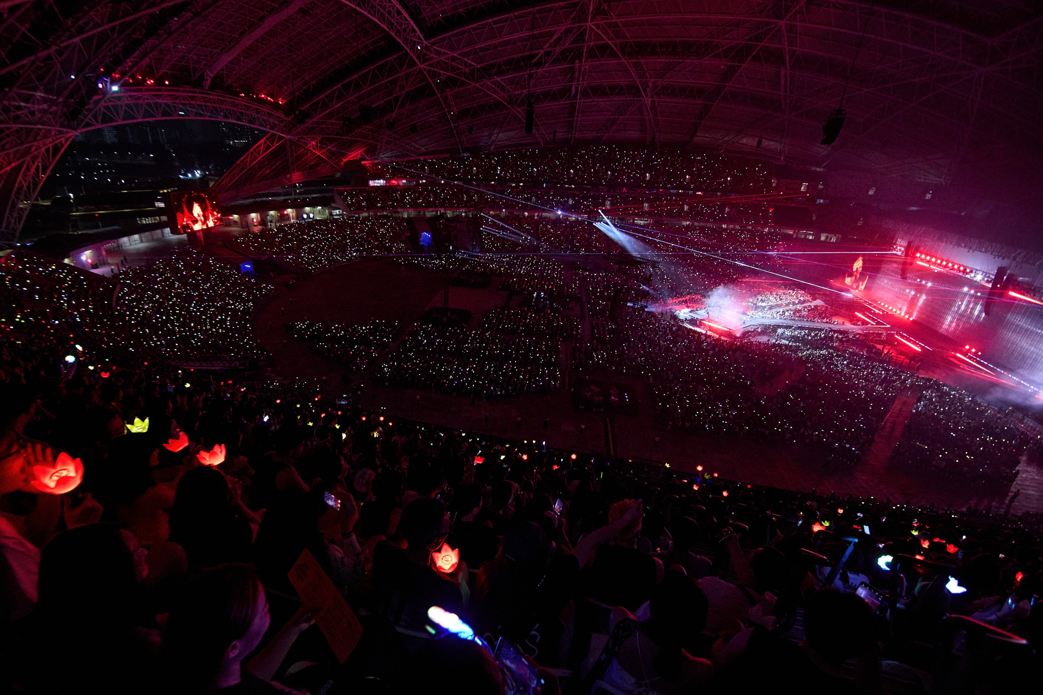 A large crowd attending a concert inside an illuminated stadium, with colorful lightsticks and laser lights creating a vibrant atmosphere.