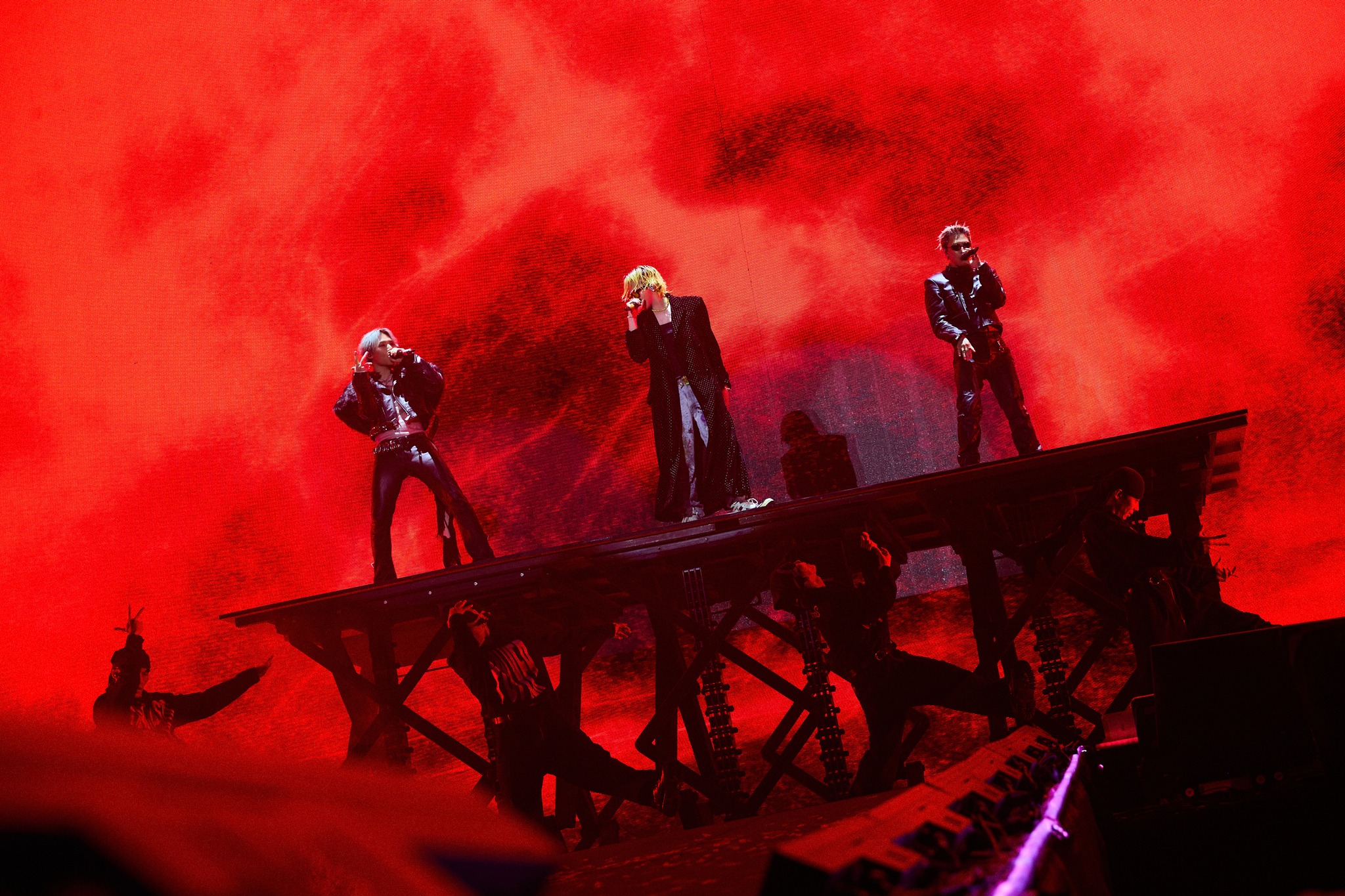 Three performers on stage during a high-energy concert, with a dramatic red background and smoke effects.