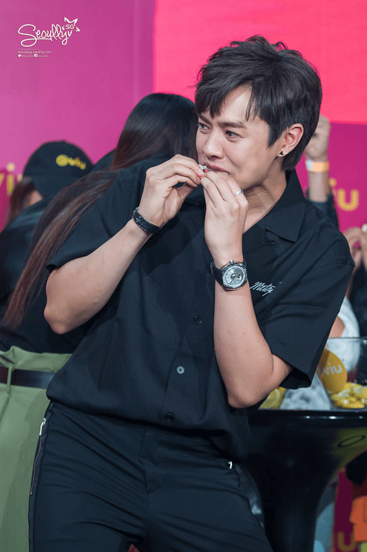 A smiling person in a black shirt playfully mimicking a chewing gesture with their hands, surrounded by a group of people in the background.