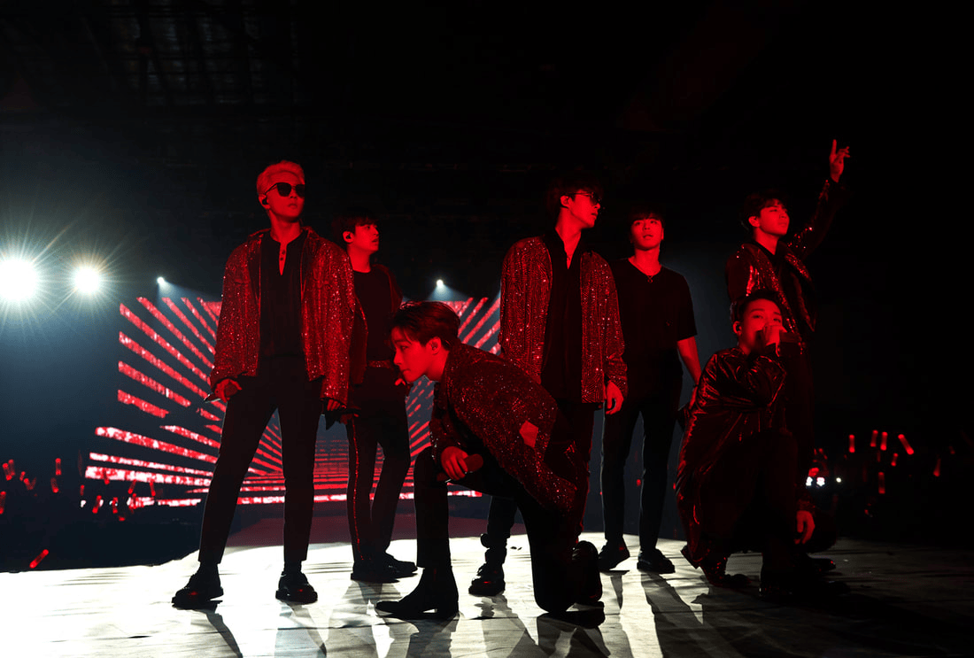 A group of seven performers on stage, dressed in black and sparkly outfits, posing dramatically under bright red lights during a concert.