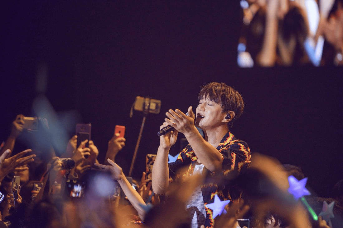 A male singer performing on stage, passionately singing into a microphone while surrounded by enthusiastic fans holding phones and light sticks.
