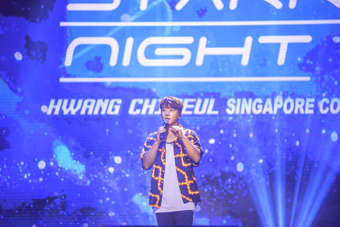 A performer speaking into a microphone on stage during an event titled 'Star Night' with a blue cosmic background and the name 'Hwang Chaeul Singapore Concert' displayed.