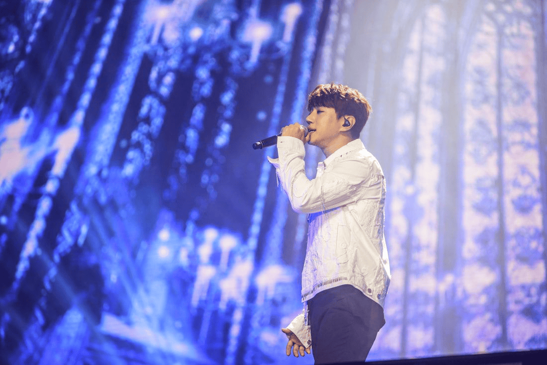 A male performer singing into a microphone on stage, wearing a white jacket, with a vibrant blue light backdrop.