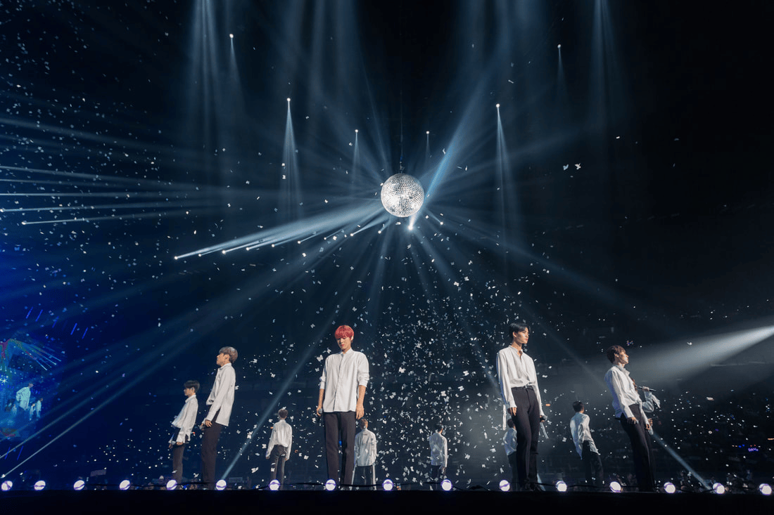 A group of performers on stage wearing white shirts, under dramatic lighting with beams and a disco ball, surrounded by falling confetti.