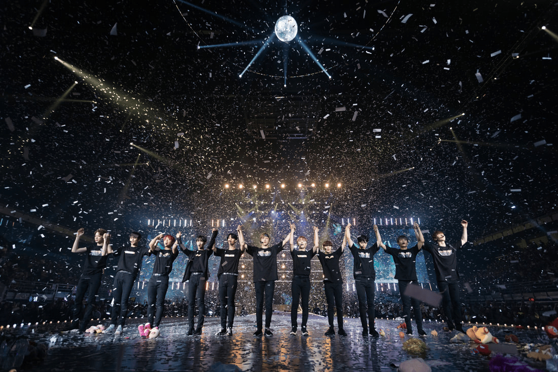 A group of performers standing on stage with their hands raised, surrounded by confetti and a spotlight. The venue is filled with an audience and colorful lighting.