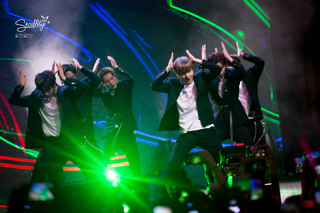 A group of male dancers performing on stage with energetic poses and synchronized movements. They are dressed in formal attire, with colorful stage lighting in the background, creating a dynamic atmosphere.