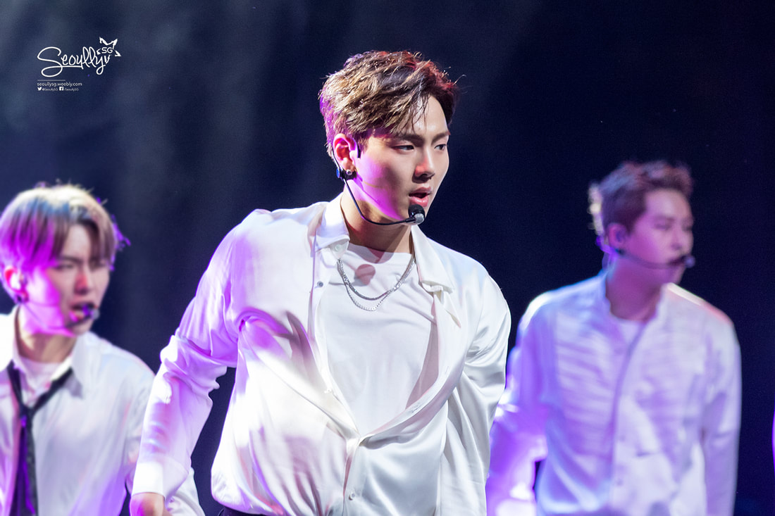 A performer on stage wearing a white shirt and necklace, focused on singing, with two other performers in the background dressed in white shirts.