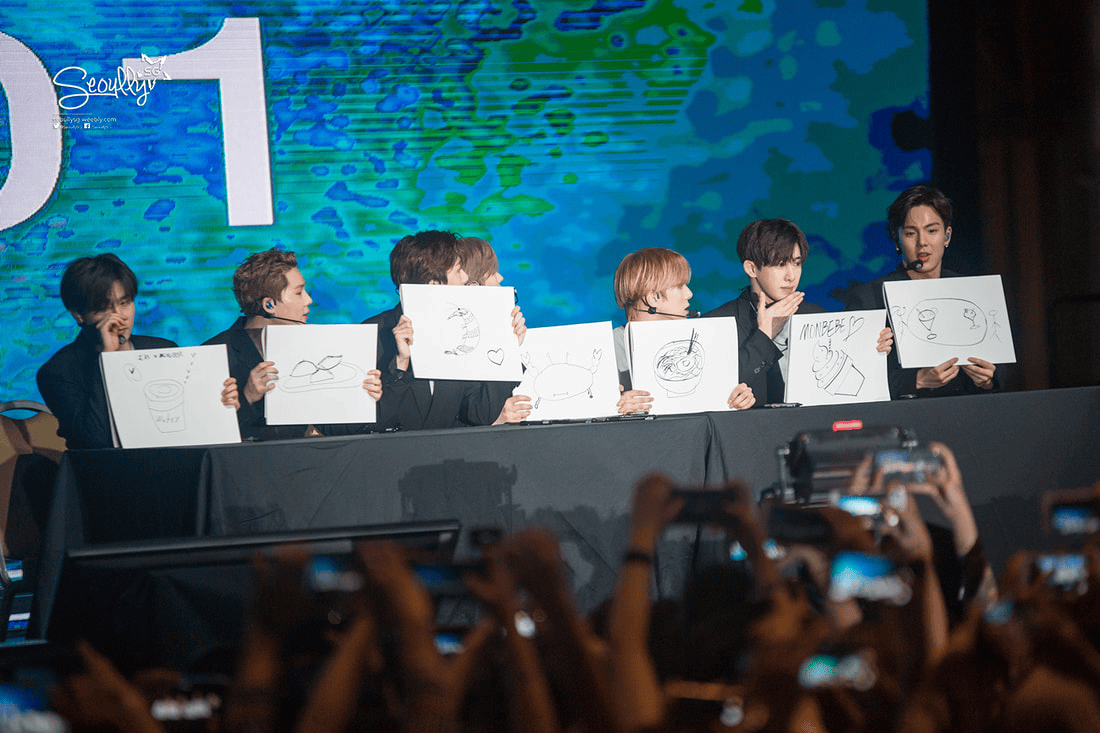 A group of seven individuals sitting behind a table, each holding up a drawing on a piece of paper during a live event, with a colorful digital background.