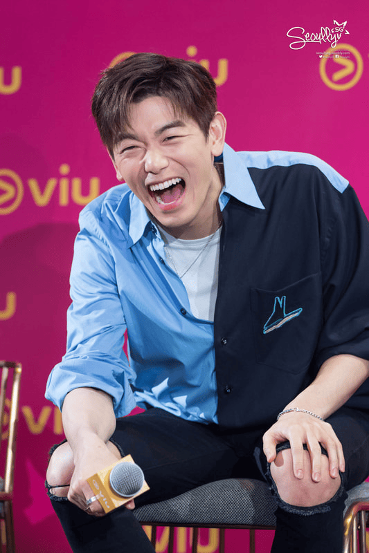 A young man with short, styled hair is laughing joyfully while sitting on a chair, wearing a half blue, half black shirt. He holds a microphone in one hand and appears to be enjoying a lively moment at an event, with a colorful background featuring the logo of a streaming service.