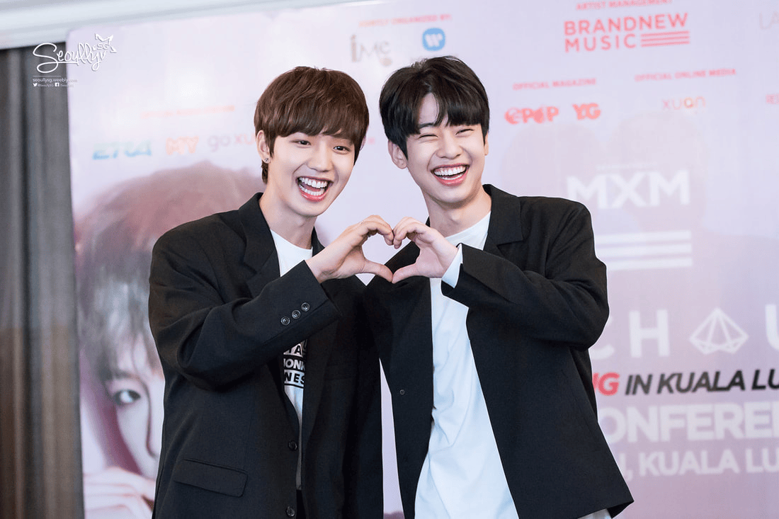 Two smiling individuals making a heart shape with their hands during an event, wearing casual outfits against a promotional backdrop.