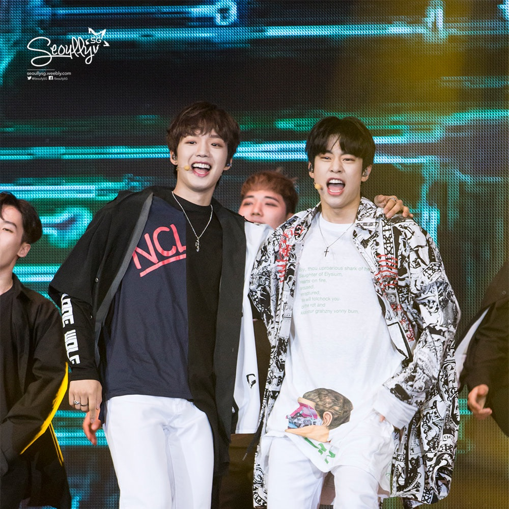 Two male performers smiling and singing on stage, wearing stylish outfits. The background features colorful stage lights.