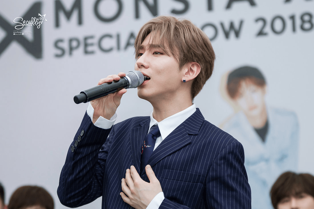 A young man in a pinstripe suit sings into a microphone, holding his chest with a sincere expression, during a special event.