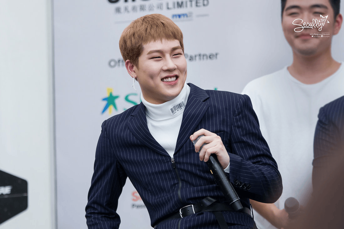 A smiling individual wearing a stylish pinstripe suit with a turtleneck, holding a microphone, appearing lively at a public event.