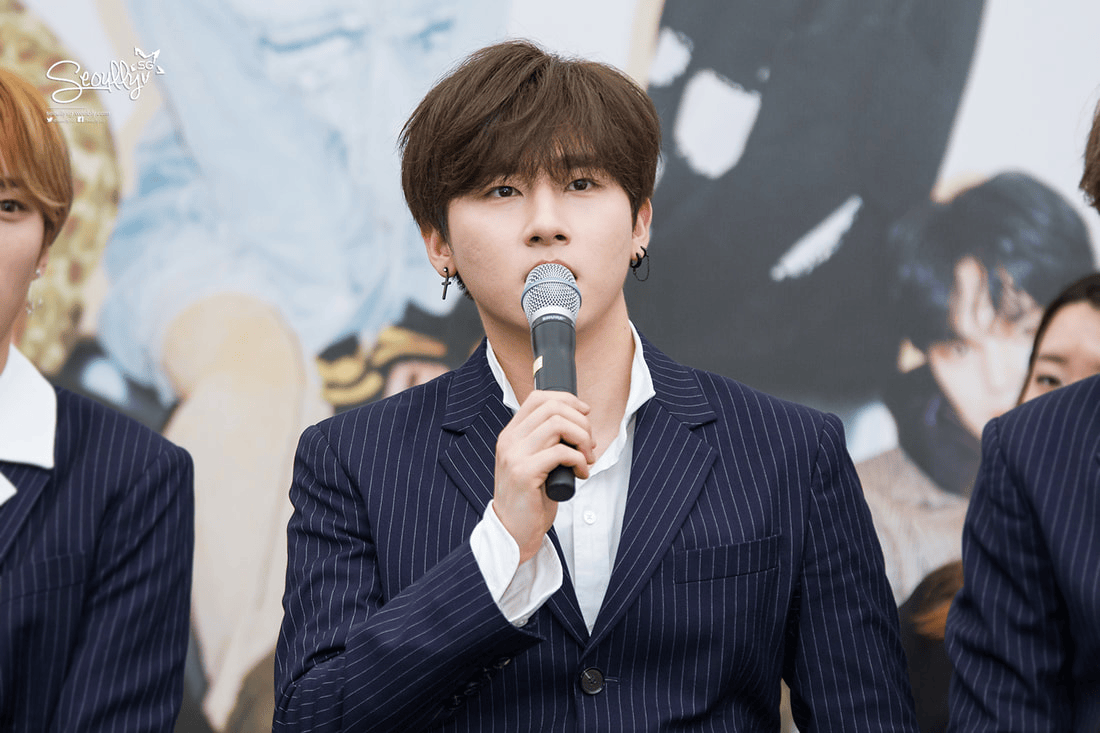 A young man with dark hair and earrings stands at a microphone, speaking during an event. He wears a pinstriped suit and looks engaged, with another person partially visible to his left.