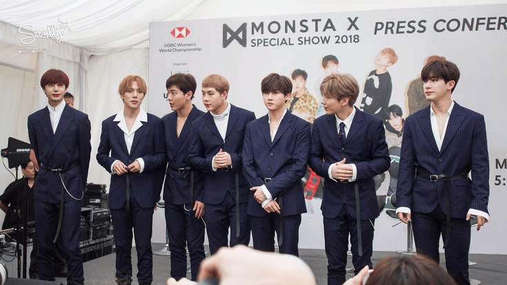 Seven members of the K-pop group Monsta X dressed in navy suits at a press conference for their 2018 special show, standing together and posing for the camera.