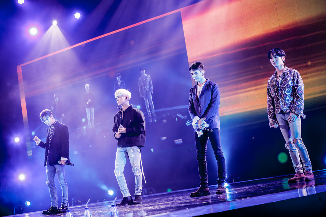 Four male performers standing on stage during a concert, illuminated by colorful lights, with a digital screen in the background.