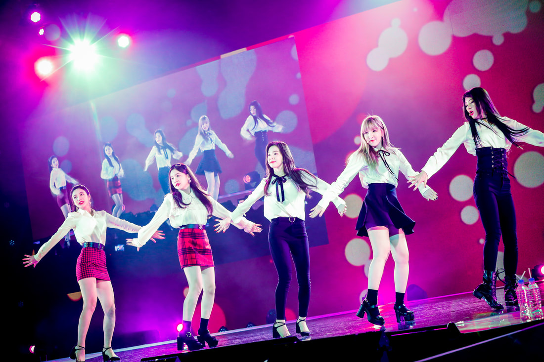 A K-pop girl group performing on stage, wearing stylish outfits, with vibrant lights and a colorful background.