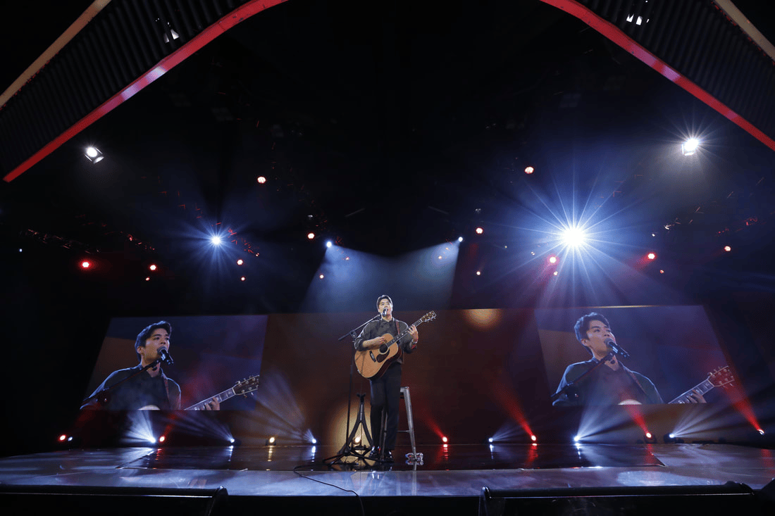 A solo musician performing on stage with an acoustic guitar, surrounded by dramatic lighting and a large backdrop screen displaying his image.
