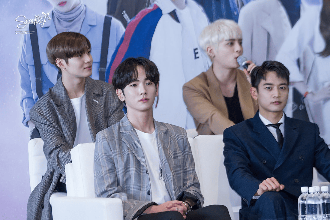 Four individuals seated at a press event, with two in the foreground looking directly at the camera while two others are positioned slightly behind them, one speaking into a microphone.