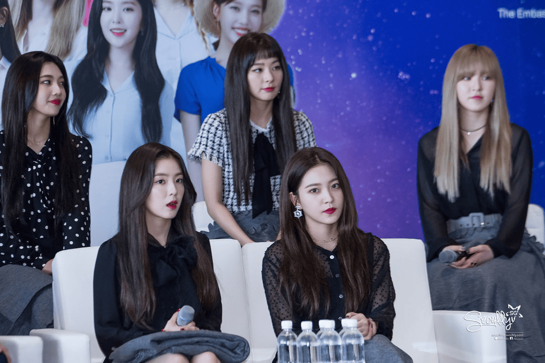 Five female performers seated during a press event, wearing stylish outfits and showing a range of expressions. In the foreground, two women are holding microphones, while bottles of water are placed on a table in front of them. A colorful backdrop featuring images of the performers is visible behind them.