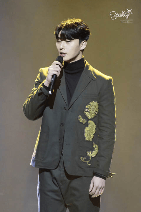 A man in a stylish green suit with intricate embroidery stands on stage holding a microphone, speaking or singing under soft lighting.