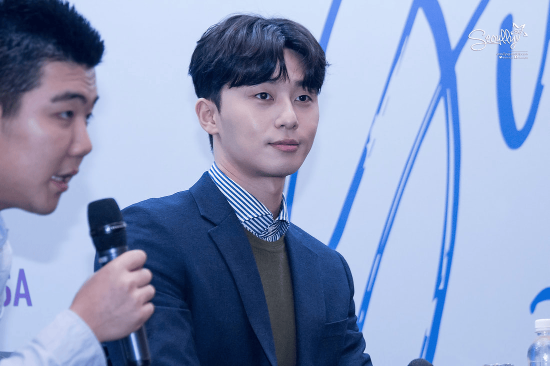 A young man in a suit listening attentively during a press conference, with another person speaking beside him.