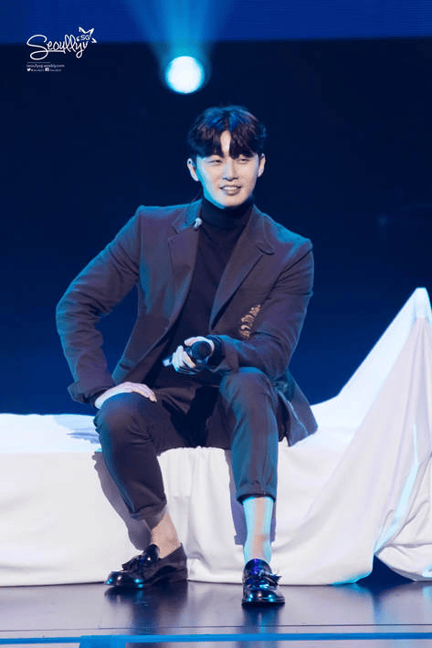 A man sitting on a stage, wearing a dark suit and smiling while holding a microphone, with a soft blue light illuminating the background.