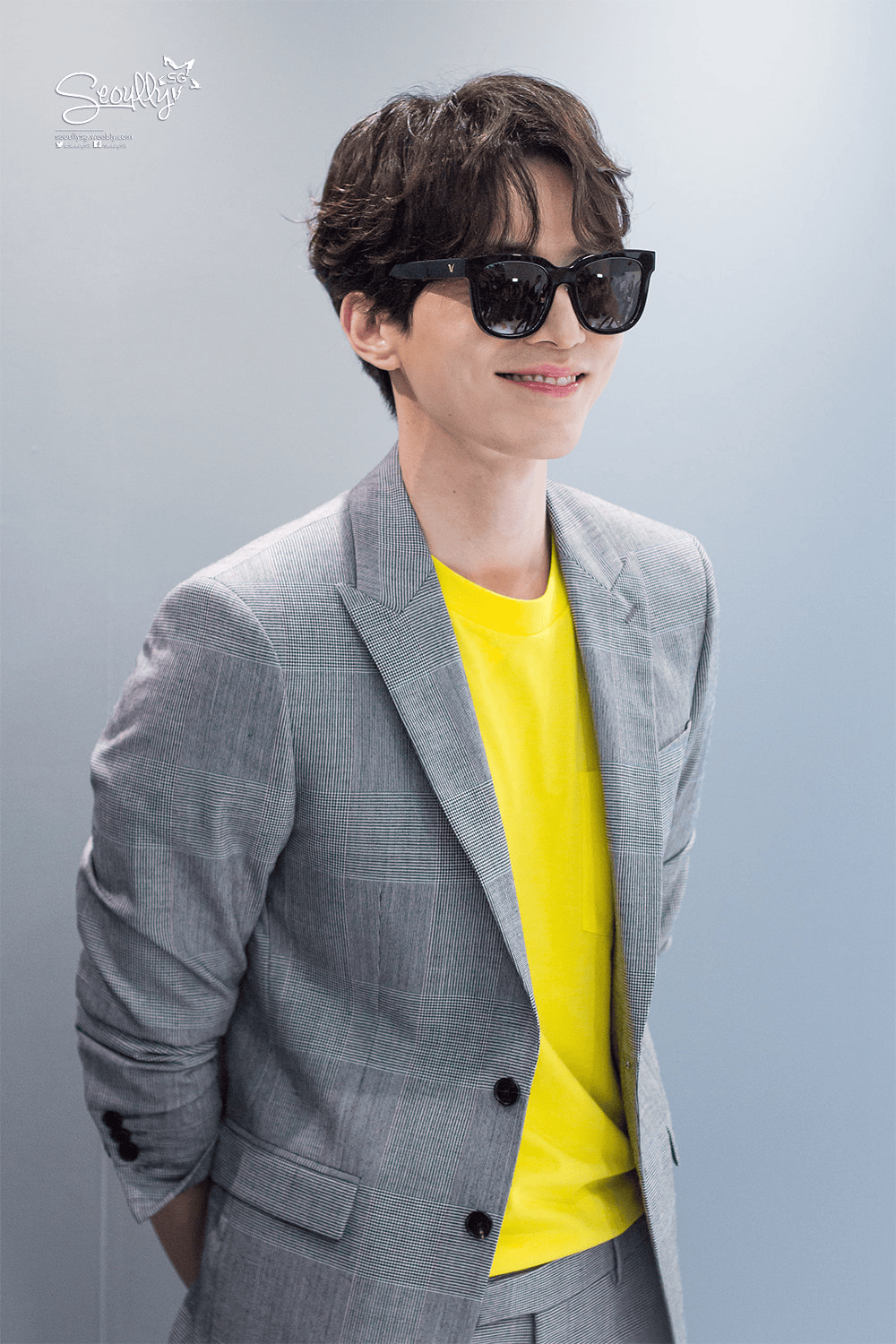 A young man in a tailored gray suit with a checkered pattern, wearing a bright yellow shirt and black sunglasses, smiling while standing against a light gray background.