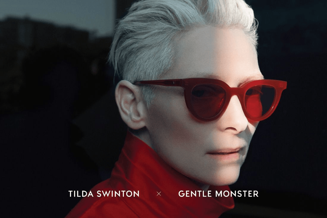 Close-up portrait of a person with short, silver hair wearing bold red sunglasses and a red outfit, set against a dark background.