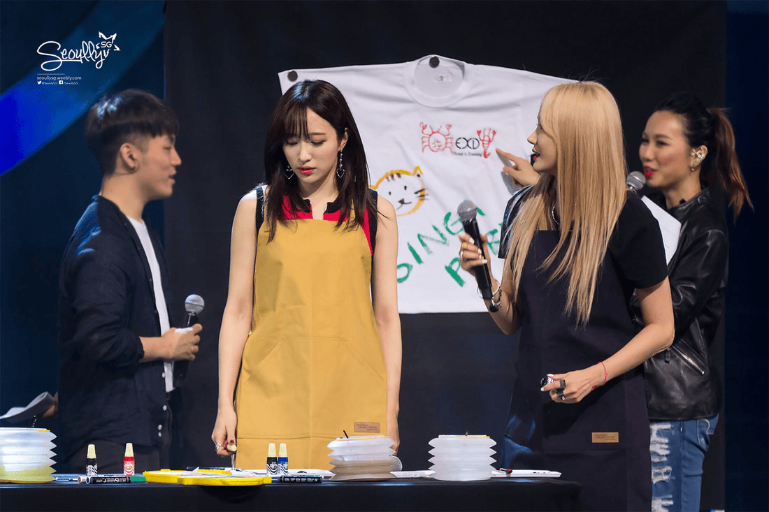 A group of individuals at a stage event, with two women wearing aprons engaged in a discussion. One woman appears to be focused, while the other gestures towards a white T-shirt hanging behind them that features artwork. A male presenter stands nearby, holding a microphone, as other colorful materials are laid out on the table.