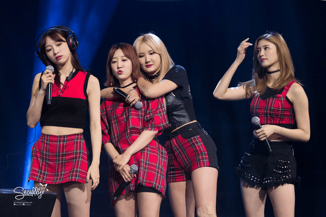 Four women in matching plaid outfits performing on stage, with one woman wearing headphones and holding a microphone.