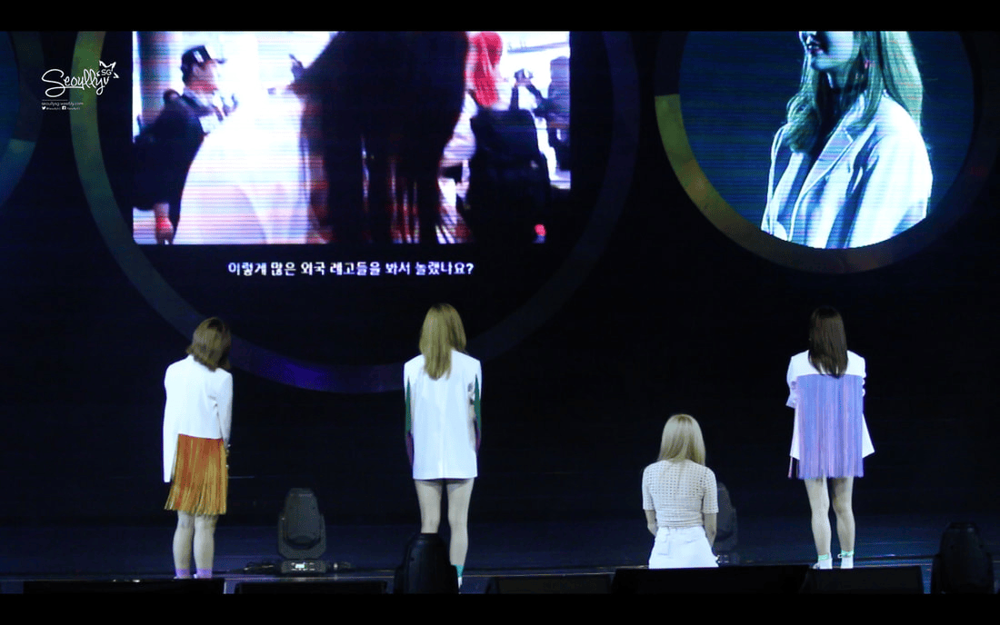 Four women standing on stage facing a large screen displaying a video, with text in Korean.