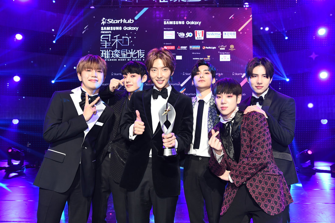 A group of six young men in suits celebrating on stage at an event, holding a trophy. They are posing and smiling in front of a brightly lit backdrop with logos.