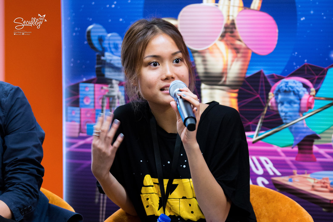 A young woman speaking into a microphone during a panel discussion, wearing a black t-shirt and gesturing with her hands, with a colorful backdrop featuring various digital art elements.