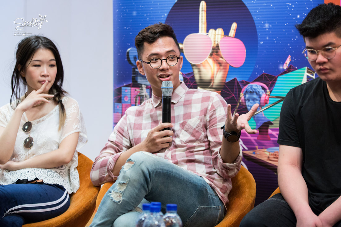 A panel discussion featuring three individuals. A man in a checkered shirt speaks into a microphone, gesturing with his hands. To his left, a woman in a white blouse and sunglasses appears thoughtful, while to his right, a man in a black shirt listens attentively. The backdrop features vibrant graphics.