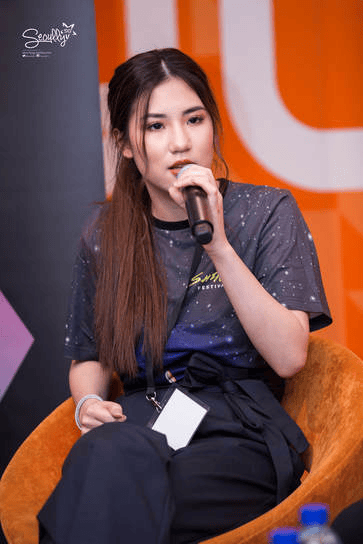 A young woman with long dark hair speaks into a microphone while seated on a chair, wearing a space-themed t-shirt and black pants at a conference.