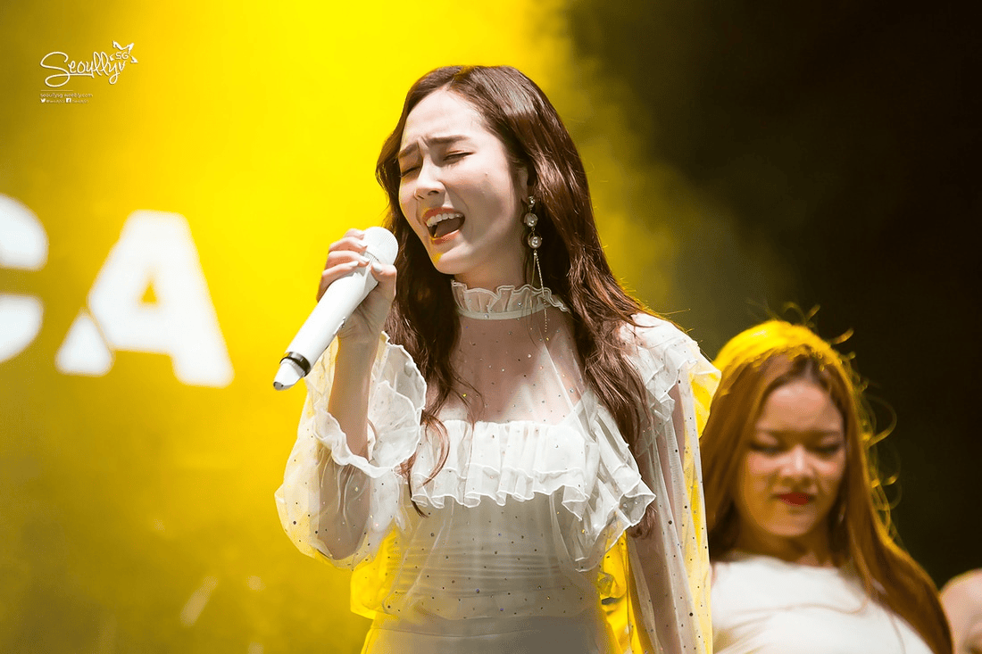 A female singer passionately performing on stage, holding a microphone, with a yellow background and soft lighting.