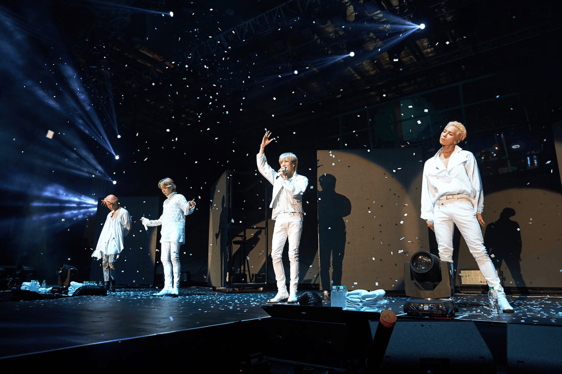 Four performers on stage in white outfits, surrounded by falling confetti, with dramatic lighting in the background.