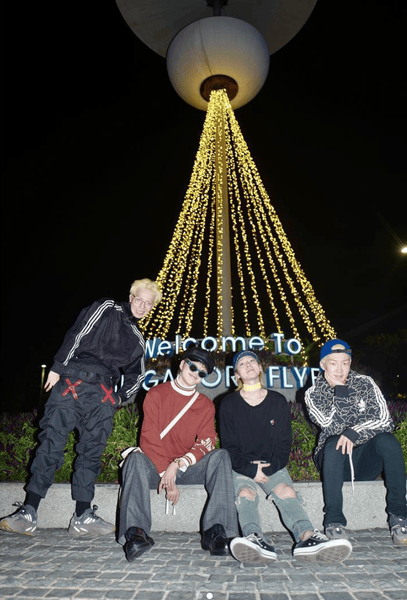 Four friends posing together at night in front of a large illuminated sign that says 'Welcome To GALLERY FLY'. The scene features decorative lights hanging down, creating a festive atmosphere.