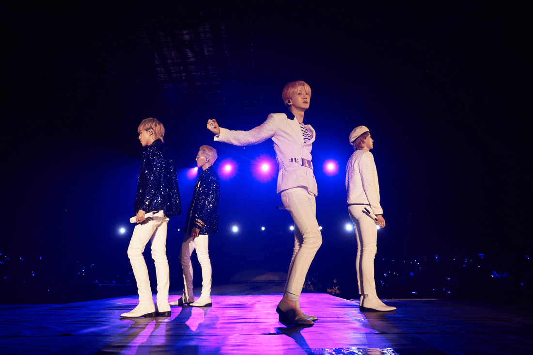Four performers on stage during a concert, dressed in sparkling jackets and white pants, with vibrant stage lights in the background.