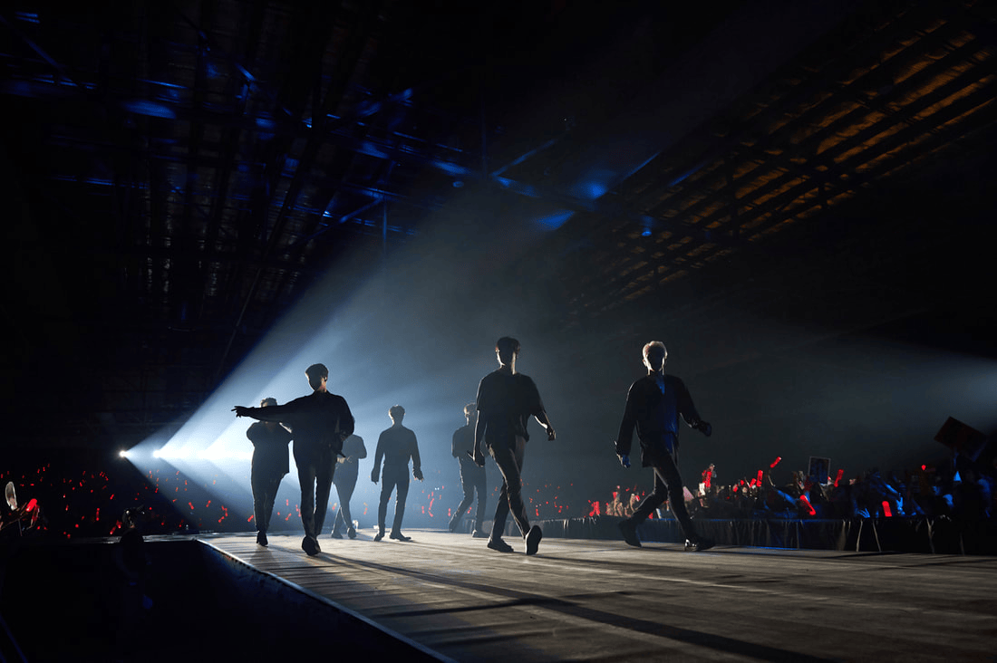 Silhouetted performers walking on stage in a dimly lit arena with colorful lights and an energetic crowd in the background.