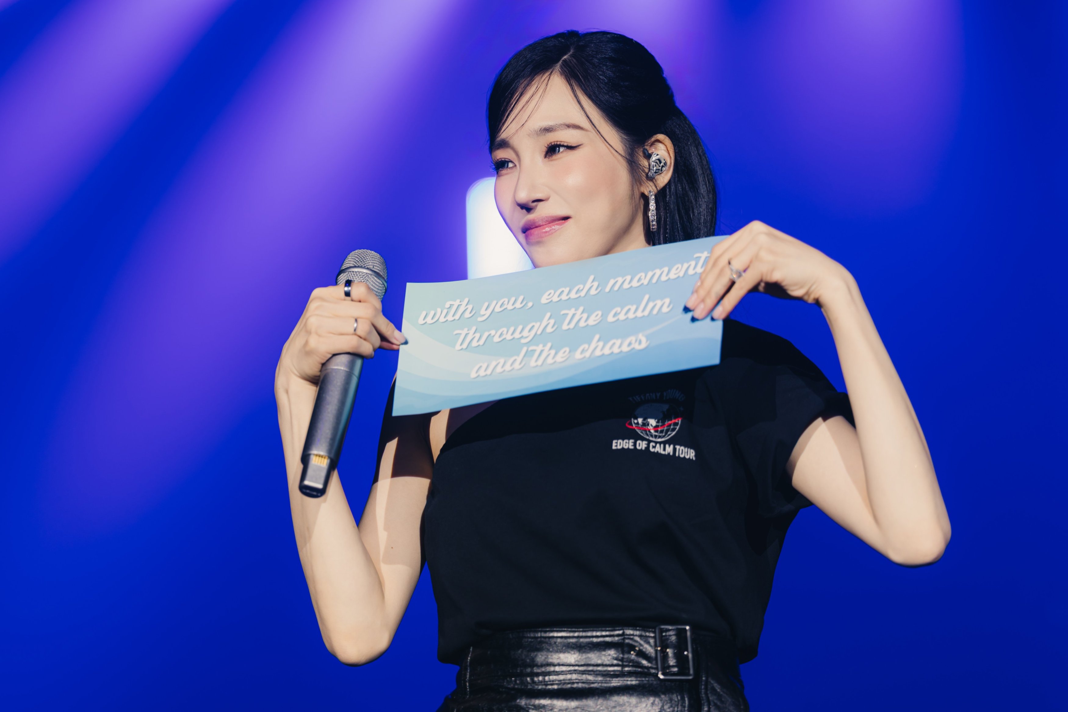 A performer holding a sign with the text 'with you, each moment through the calm and the chaos' while standing on stage with a microphone.