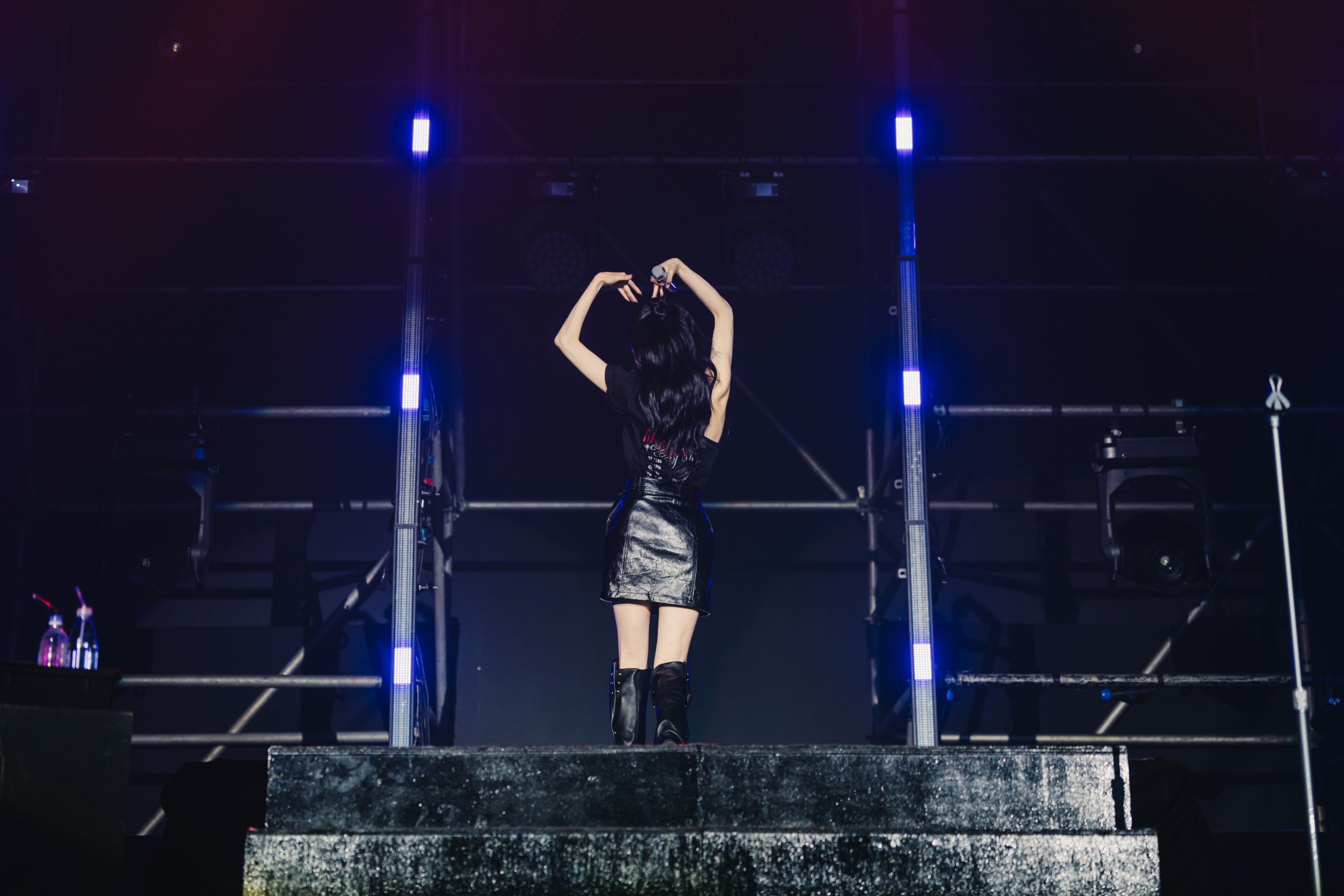 A performer on stage seen from behind, making heart shapes with their hands, wearing a black outfit and boots, illuminated by stage lights.