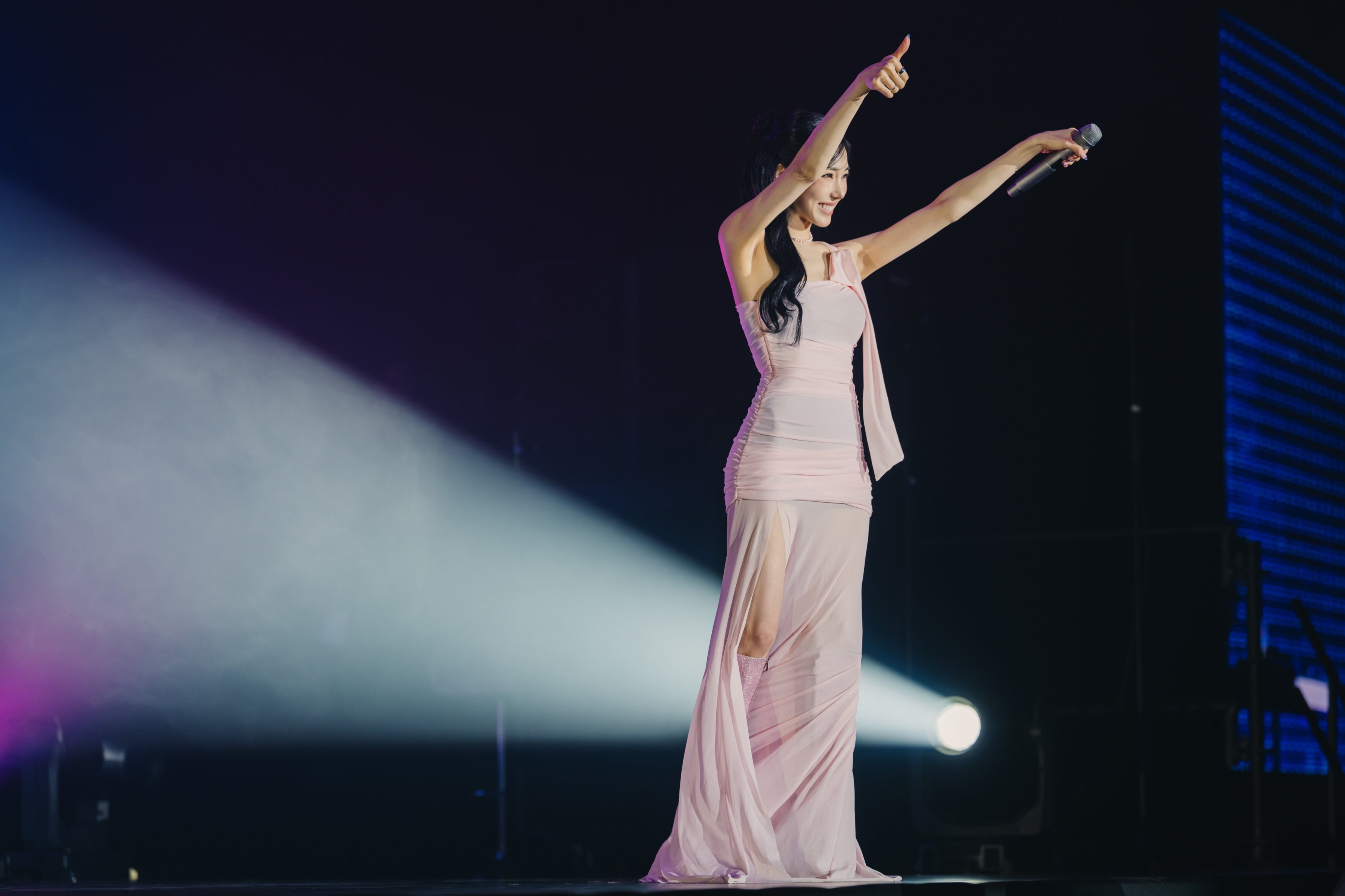 A performer in a pink dress gestures enthusiastically with one hand raised while holding a microphone in the other on stage under dramatic lighting.